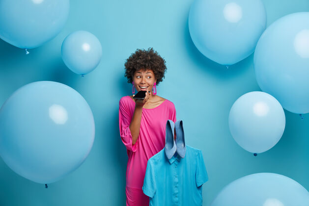 室内积极健谈的女人打电话 和朋友商量主题派对穿什么更好 拿着蓝色衬衫和鞋子 穿着粉色连衣裙 对着大氦气球在室内摆姿势手机姿势女人