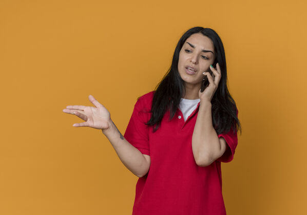 穿恼怒的年轻黑发高加索女孩穿着红衬衫在电话里说话 指着橙色墙上孤立的一面女人尖衬衫