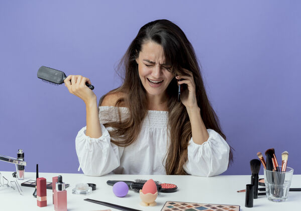 工具哭泣的美女拿着化妆工具坐在桌边拿着梳子讲电话看着紫色的墙桌子女人紫色
