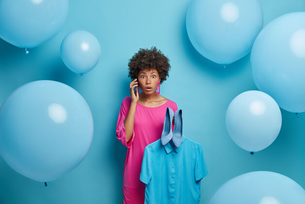 选择照片中的非洲裔美籍女子情绪激动 身穿粉色晚礼服 被节日气球包围 听到惊艳消息感到震惊 衣架上拿着衬衫和蓝色鞋子 盛装庆祝年轻风格时尚