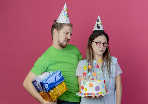 蛋糕不高兴的年轻人戴着派对帽拿着礼盒看着恼怒的年轻女孩戴着派对帽拿着生日蛋糕孤立在粉红色的墙上盒子礼物年轻