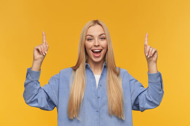 指向积极的女孩 快乐的女人 金色长发穿着蓝色衬衫人和情感的概念看着相机 手指指向复制空间 孤立在橙色背景服装表情衬衫