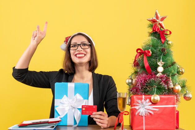 银行微笑迷人的女士穿着西装 戴着圣诞老人的帽子和眼镜 展示着礼物和银行卡 指着黄色的办公室礼物圣诞老人向上