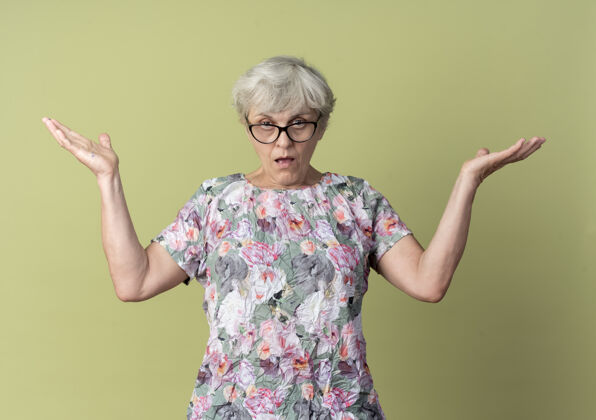 手戴着眼镜的自信的老妇人举起双手 看起来孤立无援地站在橄榄绿的墙上老女人光
