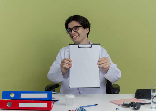 人快乐的年轻男医生 穿着医用长袍 戴着听诊器 戴着眼镜 坐在办公桌旁 带着医疗工具 看上去很孤独男性成人听诊器