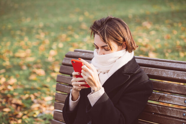 冷穿着黑色冬衣 戴着围巾 坐在公园长凳上 拿着手机的白种女人的户外画像公园举行黑发