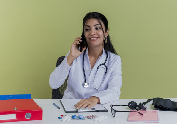坐着面带微笑的年轻女医生穿着医用长袍和听诊器坐在办公桌旁拿着医疗工具看着一旁把手放在办公桌上讲电话微笑听诊器绿色