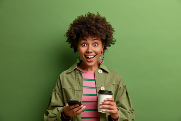 设备喜出望外的皮肤黝黑的千禧一代女孩很高兴在智能手机上收到意想不到的信息 手里拿着一杯充满活力的咖啡 穿着时髦的服装 在绿色的墙上摆姿势快乐休闲反应