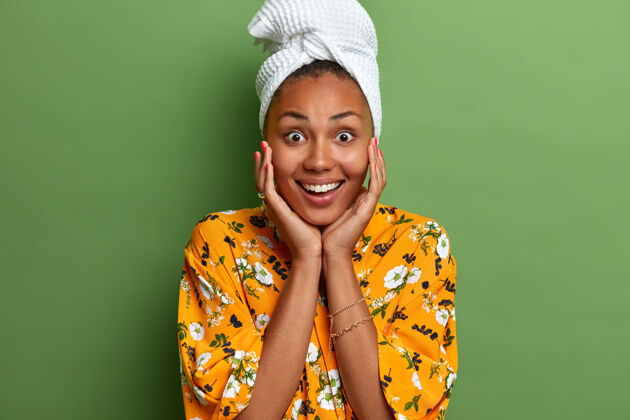 女人微笑积极的黑皮肤女人轻轻地抚摸着脸 准备淋浴后的美容程序 头上戴浴巾 黄色印花连衣裙 隔离在绿色的墙上水疗治疗外观