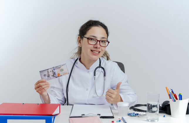 女人快乐的年轻女医生 穿着医用长袍 戴着听诊器和眼镜 坐在办公桌旁 拿着医疗工具 手里拿着钱 神情孤立地竖起大拇指快乐女性金钱