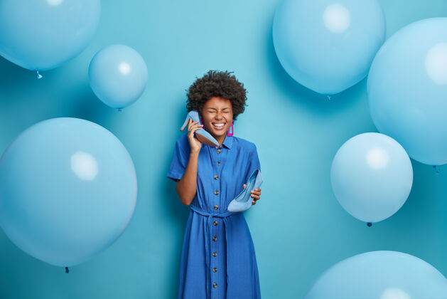 气球照片中的美国黑人妇女在迪斯科舞会上穿衣服前开怀大笑 模仿电话交谈 把高跟鞋放在耳边 孤立在蓝色的墙上年轻充气模仿