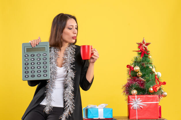 性感圣诞节的心情 微笑的美丽女士站在办公室里 拿着计算器杯在黄色的办公室里人肖像快乐
