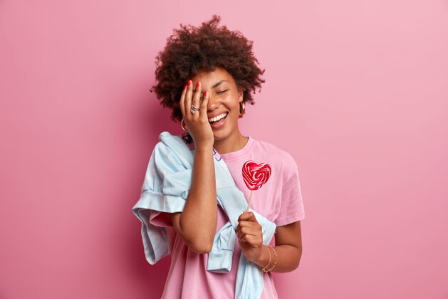 头发积极向上的非洲裔美国少女做掌心 玩得很开心 穿着休闲服 捧着心形棒棒糖 享受业余时间 对着粉色的墙壁摆姿势糖果概念毛衣牙关肖像