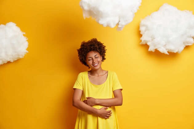 年轻快乐的非洲裔美国妇女拥抱怀孕的肚子 表达对未出生的孩子的爱 开心地微笑 享受怀孕的最后几个月 隔离在黄色的墙壁上孕妇拥抱肚子腹部等待快乐