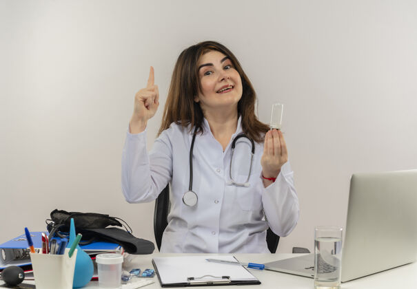 剪贴板印象深刻的中年女医生穿着医用长袍和听诊器坐在办公桌前 拿着医疗工具剪贴板和笔记本电脑拿着灯泡 竖起手指工具手指中年