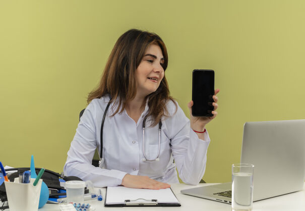 医疗高兴的中年女医生穿着医用长袍和听诊器坐在办公桌旁 拿着医疗工具和笔记本电脑 孤立地看着手机中年笔记本电脑绿色