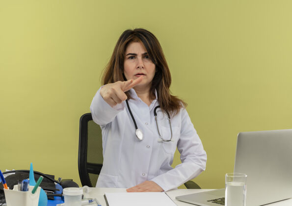 皱眉皱眉的中年女医生穿着医用长袍和听诊器坐在办公桌旁 拿着医疗工具剪贴板和笔记本电脑做手势穿着剪贴板女性