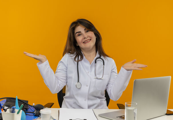 笔记本电脑微笑的中年女医生穿着医用长袍和听诊器坐在办公桌旁 带着医疗工具剪贴板和笔记本电脑 双手空空如也微笑橙色人