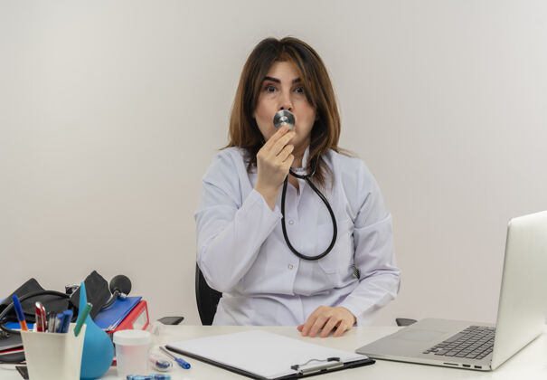 坐着印象深刻的中年女医生穿着医用长袍和听诊器坐在办公桌前用医疗工具剪贴板和笔记本电脑触摸嘴唇听诊器隔离穿着女士医疗
