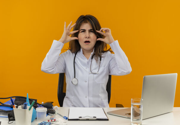 困惑困惑的中年女医生穿着医用长袍和听诊器坐在办公桌前 用医疗工具剪贴板和笔记本电脑触摸头部隔离剪贴板工具成人
