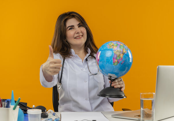 工具微笑的中年女医生穿着医用长袍和听诊器坐在办公桌旁 拿着医疗工具剪贴板和手提电脑 拿着地球仪 拇指向上伸出向上桌子医生