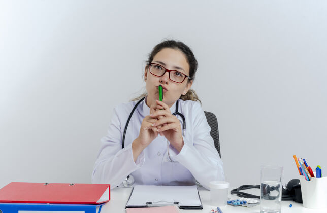 医生体贴的年轻女医生 穿着医用长袍 戴着听诊器 戴着眼镜 坐在办公桌前 拿着医疗工具 用笔抚摸着嘴唇 孤零零地抬头看医生桌子人