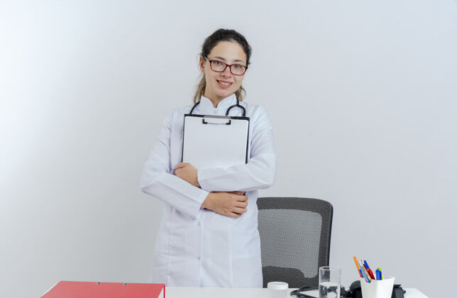 长袍面带微笑的年轻女医生穿着医用长袍 戴着听诊器和眼镜 站在桌子后面 带着医疗工具 看上去与世隔绝桌子站着眼镜