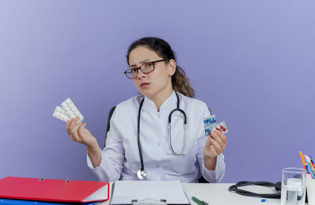 抱着愁眉苦脸的年轻女医生穿着医用长袍 听诊器坐在办公桌旁 手里拿着医疗工具 手里拿着药片 看上去与世隔绝药片医生成人