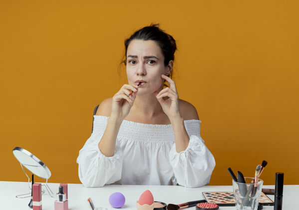 桌子焦急美丽的女孩坐在桌子旁用化妆工具咬着隔离在橙色墙上的化妆刷化妆焦虑橘子