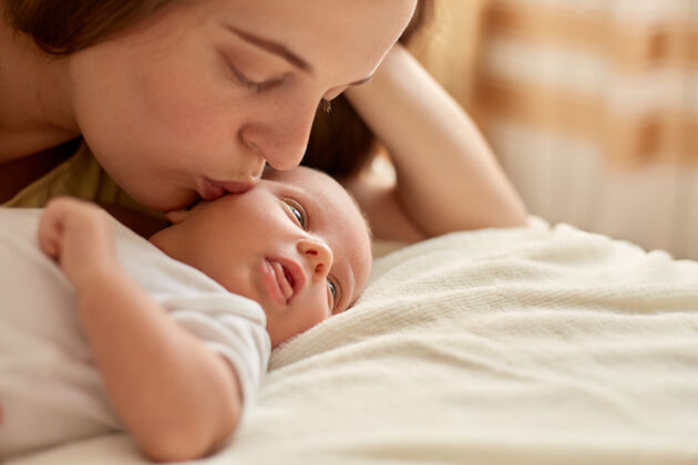女儿母亲和她的新生婴儿一起躺在床上的毯子上快乐的母亲亲吻和拥抱婴儿 孩子看着别处 学习外面的东西母性和亲子关系家庭温柔爱