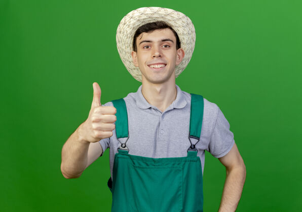 园艺微笑着的年轻男园丁戴着园艺帽 竖起大拇指孤立地站在绿色背景上 留着复制空间穿年轻绿色