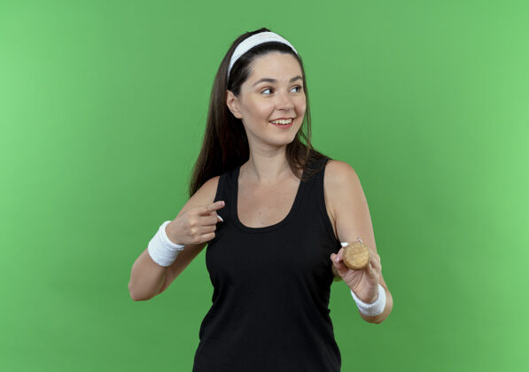 站着戴着头巾的年轻健身女士拿着棒球棒 面带微笑 手指着站在绿色背景上的一边健身棒球抱着