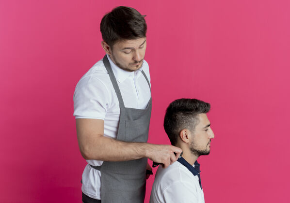 剪穿着围裙的理发师在粉色背景上用客户满意的理发机剪头发围裙头发客户