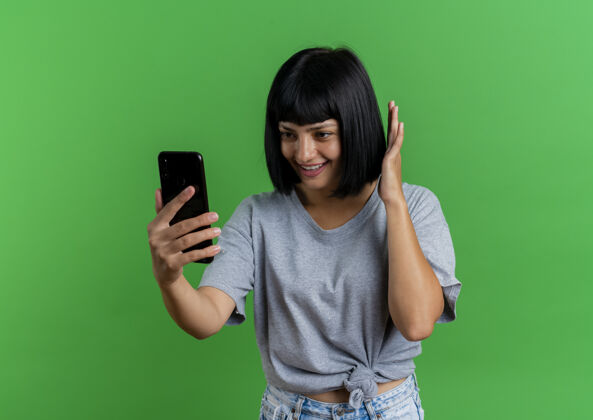 复制年轻的黑发白人女孩微笑着举手 看着绿色背景上的手机和复印空间黑发年轻绿色
