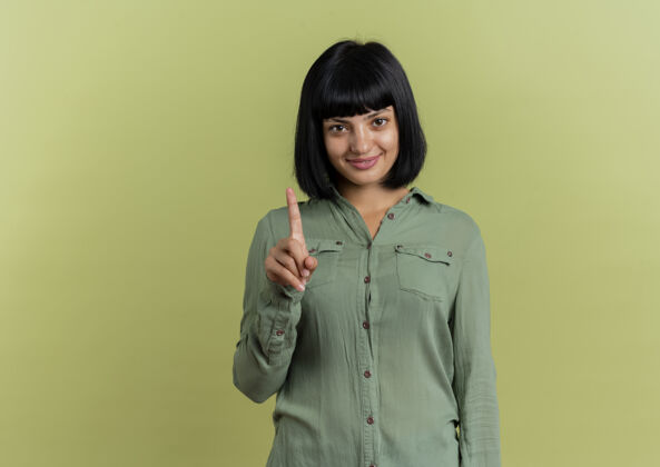 年轻微笑的年轻黑发白人女孩显示食指在橄榄绿的背景与复制空间孤立的相机复制手指空间