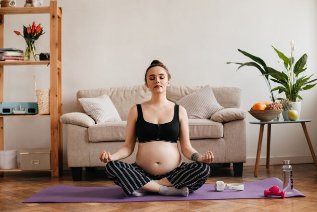 父母穿着裤子和瑜伽上衣的孕妇在米色沙发旁冥想坐着休息室内