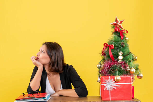 微笑坐在黄色办公室装饰圣诞树旁的桌子旁看东西的年轻女子东西肖像圣诞节