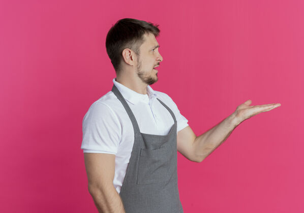 争论穿着围裙的理发师站在粉色背景上 伸出手臂 一边看一边询问或争论理发师围裙问
