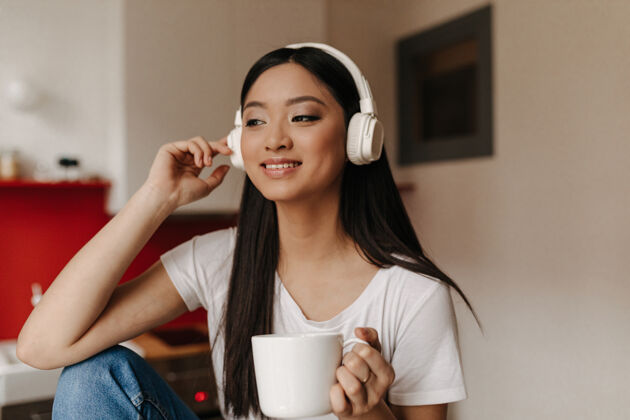 休闲穿着白色t恤和耳机的美女边喝茶边听音乐耳机房子音乐