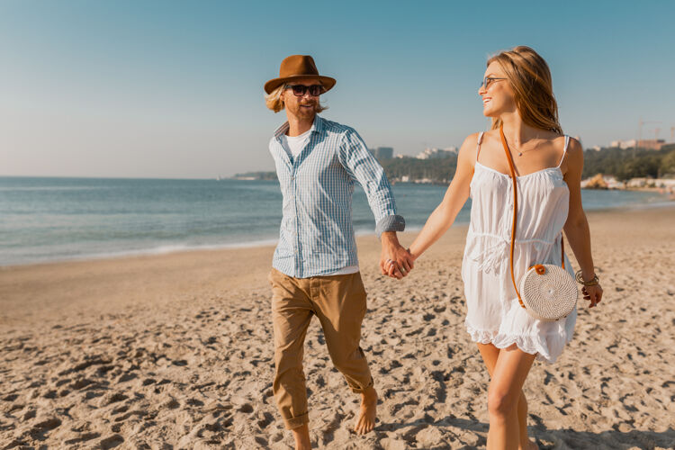 旅游者年轻迷人的微笑着戴着帽子的快乐男人和穿着白裙子的金发女人一起在沙滩上奔跑旅行浪漫男朋友