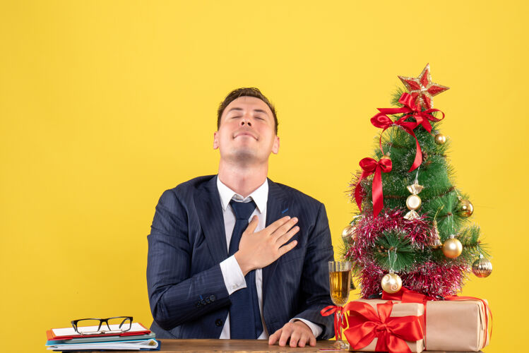 礼物正面图幸福的男人把手放在胸前 坐在圣诞树旁的桌子旁 黄色背景的礼物坐着生意人生意