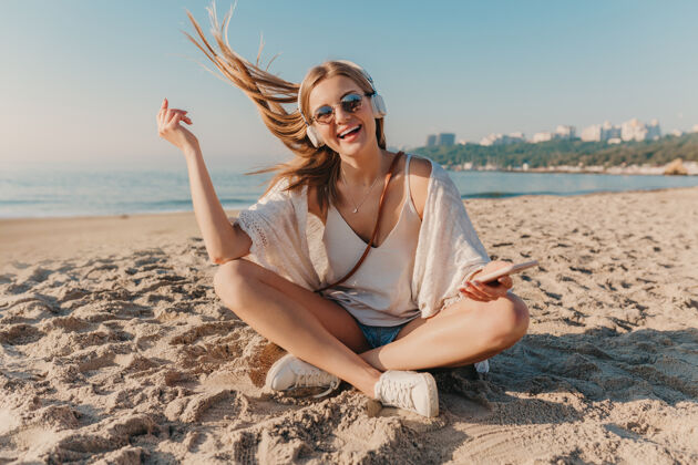 沙滩年轻迷人的金发微笑的女人坐在沙滩上 戴着耳机 以积极的心情听音乐音乐海洋海洋