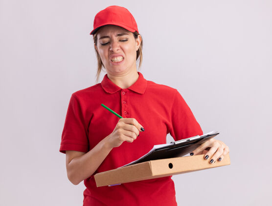 制服穿着制服的年轻漂亮的送货员拿着铅笔和剪贴簿放在隔离在白墙上的比萨饼盒上人不愉快剪贴板