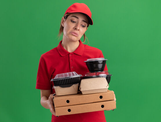 站穿着制服的悲伤的年轻漂亮的女送货员拿着纸食品包装和比萨饼盒上的容器隔离在绿色的墙上纸食物表情