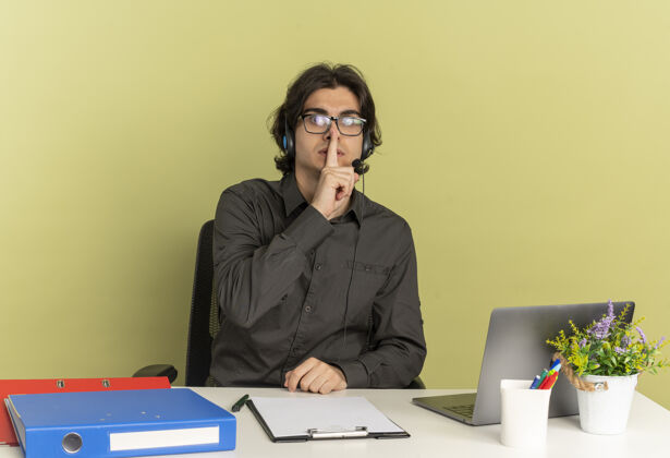眼镜年轻焦急的上班族戴着耳机戴着眼镜坐在办公桌旁 拿着办公工具用笔记本电脑把手指放在嘴上 在绿色的背景和复印空间里默不作声嘴使用工人