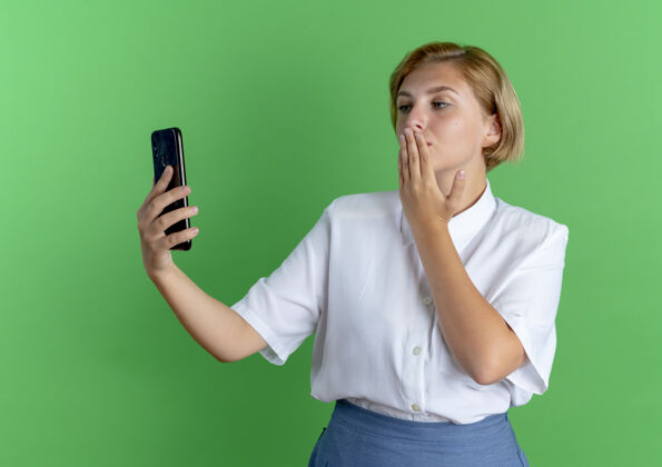 手年轻自信的俄罗斯金发女孩拿着手机 看着手机把手放在嘴上 在绿色背景上隔离 留有复印空间年轻握着女孩