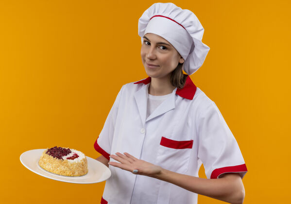 拿着身着厨师制服的年轻高加索厨师女孩高兴地拿着并指着盘子上的蛋糕 蛋糕被隔离在橙色的背景上 还有复制空间厨师蛋糕烹饪