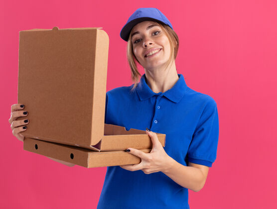 人微笑着的年轻漂亮的女送货员穿着制服 拿着隔离在粉红色墙上的比萨饼盒站着姿势披萨