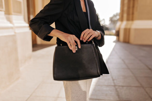 女人穿着黑色夹克和白色裤子的女人在外面拿着黑色的手提包穿着时髦衣服的女人在户外摆着时髦的手提包台阶步行街道