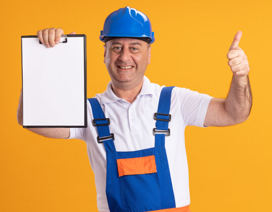 保持身着制服的成年建筑工人微笑着竖起大拇指 把剪贴板孤立地放在橙色的墙上衣服微笑姿势